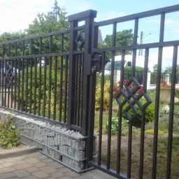 Nowoczesne ogrodzenie z metalowymi przęsłami i gabionowymi słupkami. Elementy dekoracyjne w kształcie rombów. Widoczny fragment chodnika z kostki brukowej i zieleń w tle.