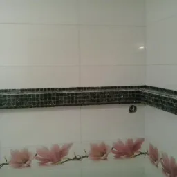 Półka z mozaiki w rogu łazienki, białe płytki z motywem kwiatowym u dołu, widoczne niedociągnięcia w fugowaniu. Ujęcie z bliska.