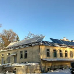 Panele słoneczne zamontowane na dachu budynku w zimowej scenerii, widoczne drzewa bez liści i niebieskie niebo, w prawym dolnym rogu kontener z logotypem firmy.