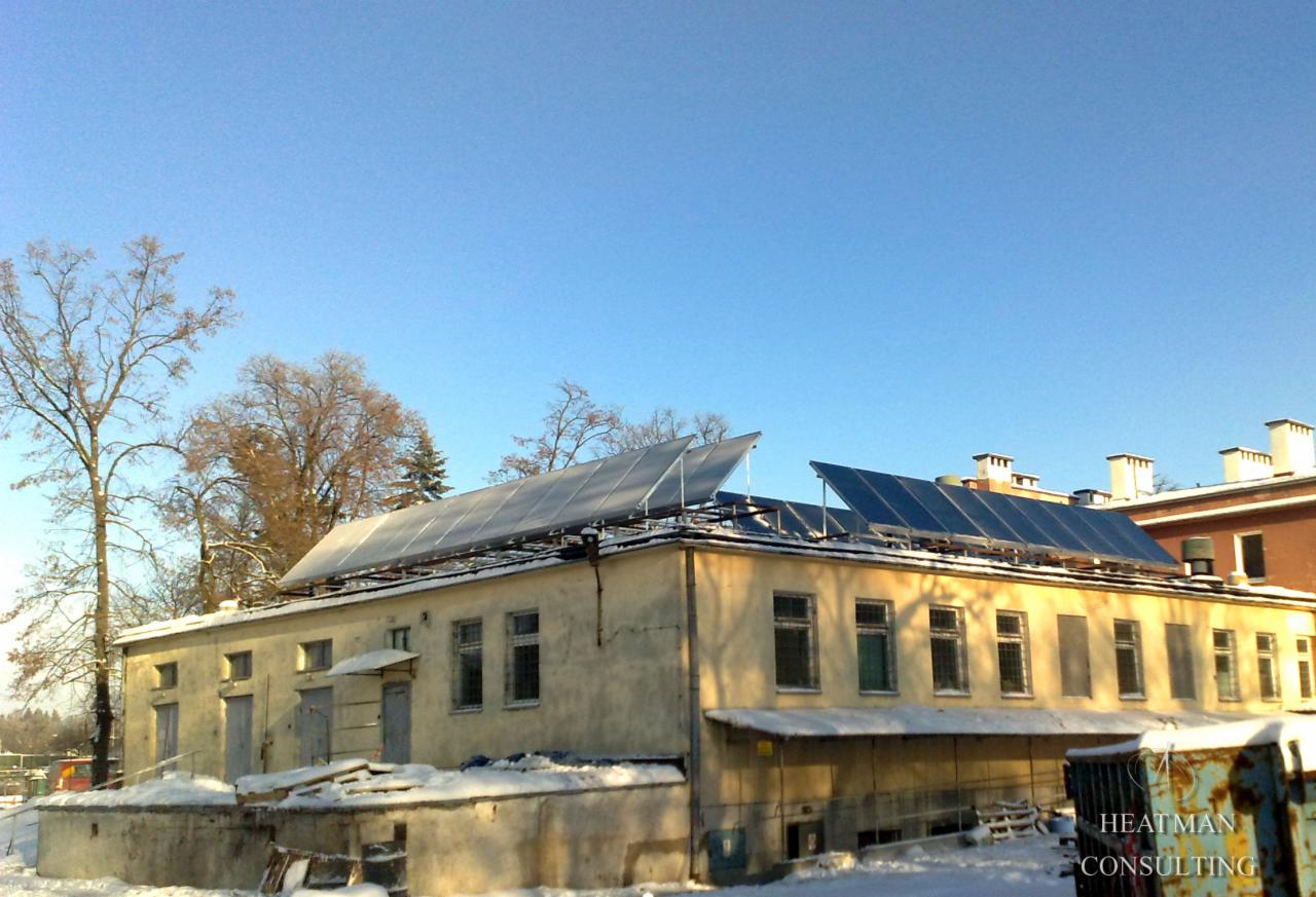 Panele słoneczne zamontowane na dachu budynku w zimowej scenerii, widoczne drzewa bez liści i niebieskie niebo, w prawym dolnym rogu kontener z logotypem firmy.