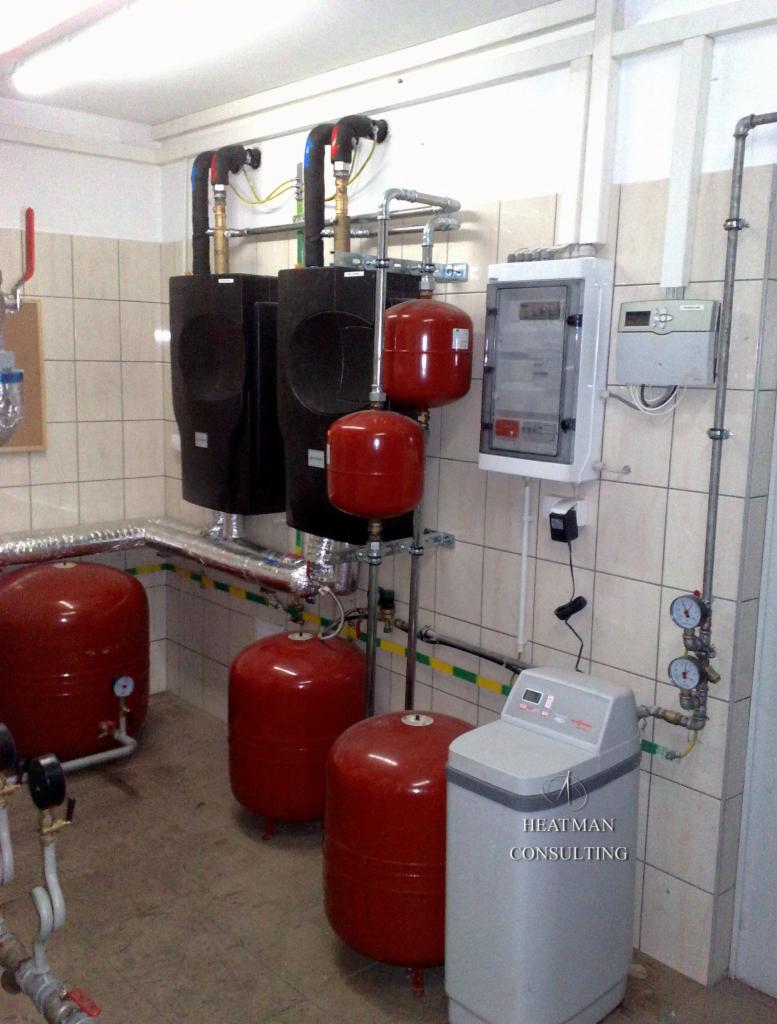 Kotłownia z dwoma czarnymi kotłami, kilkoma czerwonymi zbiornikami ciśnieniowymi, szarą stacją uzdatniania wody i rozdzielnią elektryczną na ścianie z beżowych płytek.