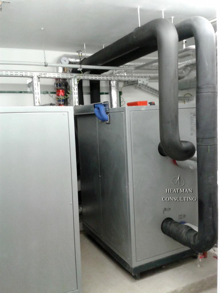 Pompa ciepła w kotłowni z izolowanymi rurami i logo Heatman Consulting na obudowie.