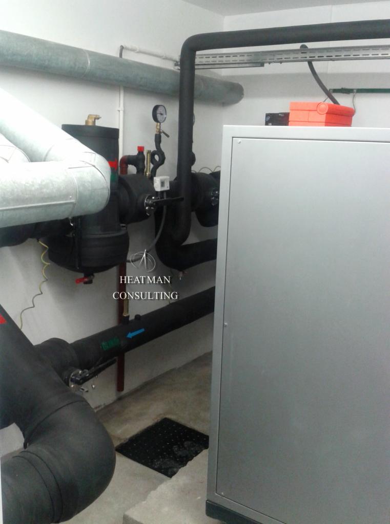 Węzeł cieplny z czarnymi rurami izolowanymi termicznie, manometrem, zaworami i szarym panelem obudowy, w pomieszczeniu technicznym.