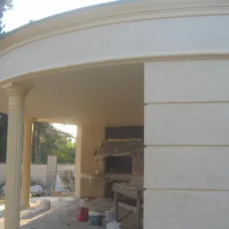 Zadaszona altana z kolumnami w stylu klasycznym. Widoczna faktura ściany i elementy dekoracyjne. W tle zieleń drzew. Na podłodze wiaderka z farbą.