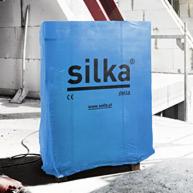 Niebieska folia ochronna z logo Silka na palecie bloczków wapienno-piaskowych, na tle surowej ściany w trakcie budowy. Widoczne fragmenty rusztowania.