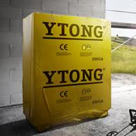 Żółta paleta bloczków Ytong na budowie, zabezpieczona folią. Widoczne logo Ytong i oznaczenia CE. W tle fragment okna i zieleń.
