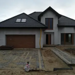 Surowy, dwukondygnacyjny dom z ciemną dachówką i drewnianą bramą garażową w trakcie wykończenia, z ułożoną częściowo kostką brukową na podjeździe i żółtymi rurami spustowymi.