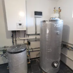 Nowa instalacja grzewcza w kotłowni: wiszący biały piec Yutaki obok dwóch srebrnych zbiorników owiniętych folią ochronną, z widocznymi rurami i zaworami.