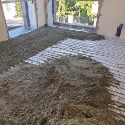 Nierównomiernie rozłożona, wilgotna masa jastrychowa na instalacji ogrzewania podłogowego w surowym pomieszczeniu z widokiem na ogród.