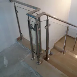 Szczegółowe ujęcie schodów z zamontowaną balustradą ze stali nierdzewnej i szkła, widoczne mocowania szkła do słupków, surowy beton na posadzce.