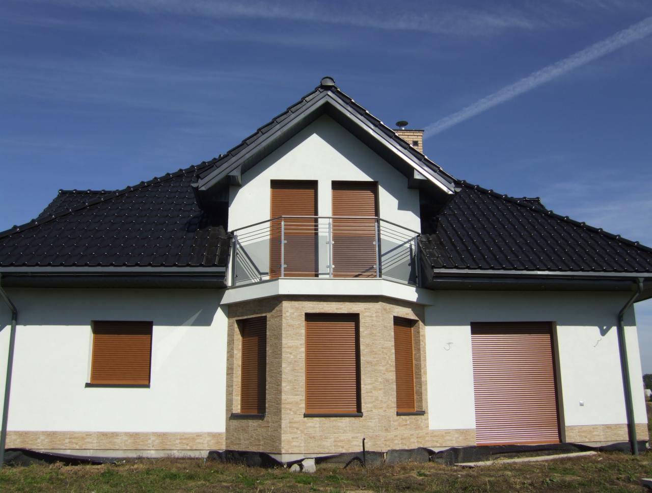 Nowoczesny balkon z szklaną balustradą w domu jednorodzinnym z brązowymi roletami zewnętrznymi i czarnym dachem na tle błękitnego nieba.