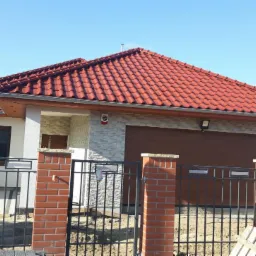 Nowoczesny dom jednorodzinny z czerwoną dachówką, elewacją z jasnego kamienia i brązową bramą garażową, widoczny zza czarnego, metalowego ogrodzenia z ceglanymi słupkami. Na pierwszym planie...