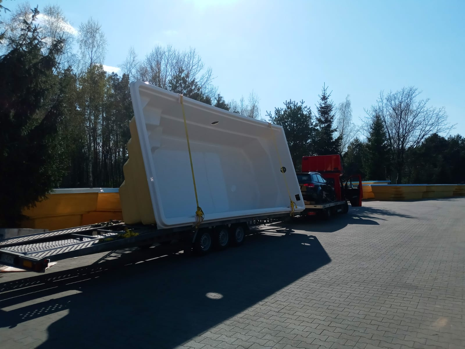 Transport otwartego, białego basenu z żółtymi wzmocnieniami narożnymi na niskopodwoziowej przyczepie, z samochodem osobowym załadowanym na czerwoną lawetę w tle, na brukowanym placu w słoneczny dzień.