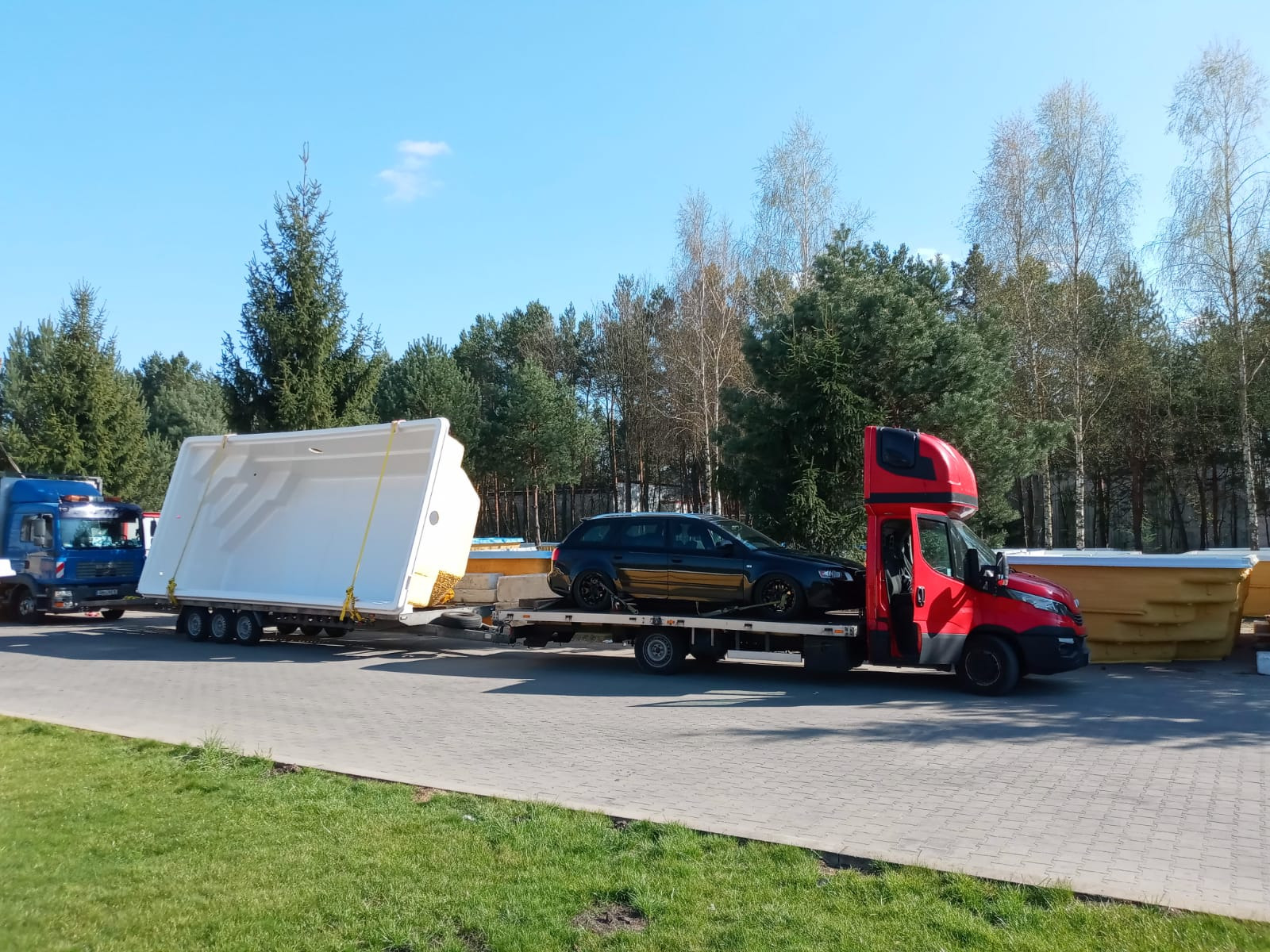 Czerwona laweta przewozi czarne kombi i biały, prostokątny basen z żółtymi pasami mocującymi na tle lasu i niebieskiego nieba.