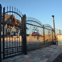 Otwarta, czarna, metalowa brama wjazdowa z dekoracyjnymi elementami w kształcie wici roślinnych, na posesję z domem o brązowym dachu i czerwonym samochodem w tle.