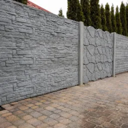 Szary betonowy płot z różnymi fakturami: imitacja kamienia, drewna i dekoracyjny ornament, widok z poziomu kostki brukowej.