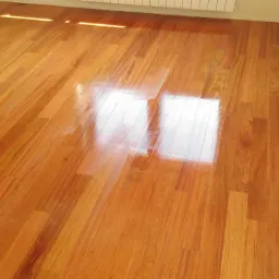 Błyszcząca, świeżo polakierowana podłoga parkietowa z widocznym odbiciem światła słonecznego, ułożona w pomieszczeniu z jasnymi ścianami i grzejnikiem.