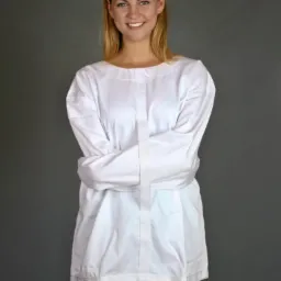 Uśmiechnięta blondynka w białej bluzie z długimi rękawami skrzyżowanymi na piersiach, na tle szarego studia.