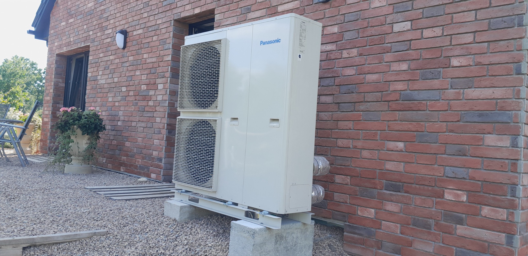 Biała pompa ciepła Panasonic zamontowana na betonowych blokach przy ceglanym budynku. Widoczne przewody i wentylatory. Zewnętrzna instalacja systemu grzewczego.
