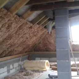 Poddasze z widoczną izolacją z wełny mineralnej w przestrzeni między krokwiami, elementy konstrukcji drewnianej i murowany komin. Widoczne okna dachowe.