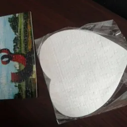 Dwa puzzle: prostokątny z nadrukiem abstrakcyjnej rzeźby i biały w kształcie serca, zapakowany w folię, leżą na ciemnym biurku.