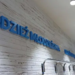 Niebieski napis 'Odzież Młodzieżowa' wykonany z liter 3D na ścianie z białej cegły w sklepie w Suwałkach, widok z boku na wieszaki na ubrania.