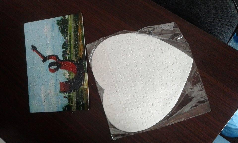 Dwa puzzle: prostokątny z nadrukiem abstrakcyjnej rzeźby i biały w kształcie serca, zapakowany w folię, leżą na ciemnym biurku.