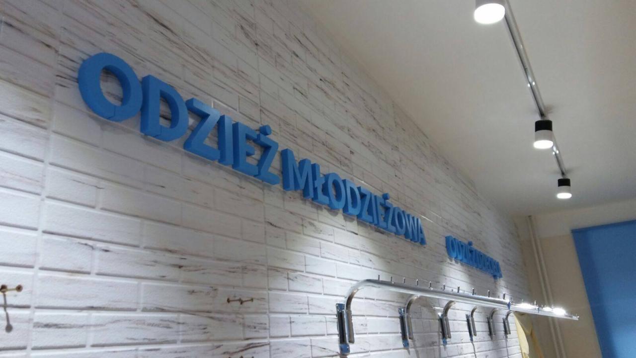 Niebieski napis 'Odzież Młodzieżowa' wykonany z liter 3D na ścianie z białej cegły w sklepie w Suwałkach, widok z boku na wieszaki na ubrania.