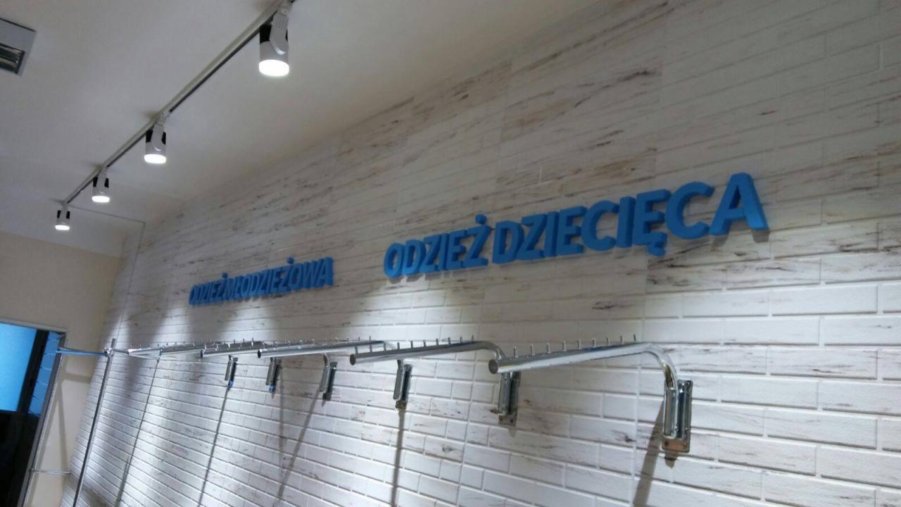 Wnętrze sklepu odzieżowego: ściana z białej cegły z niebieskim napisem 'Odzież Dziecięca' i 'Odzież Młodzieżowa', oświetlenie punktowe i metalowe wieszaki na ubrania.