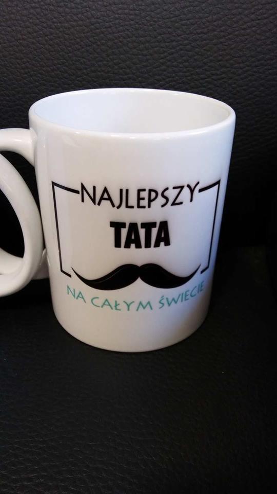 Biały kubek z czarnym nadrukiem 'Najlepszy Tata na całym świecie' i wąsami na ciemnym tle.
