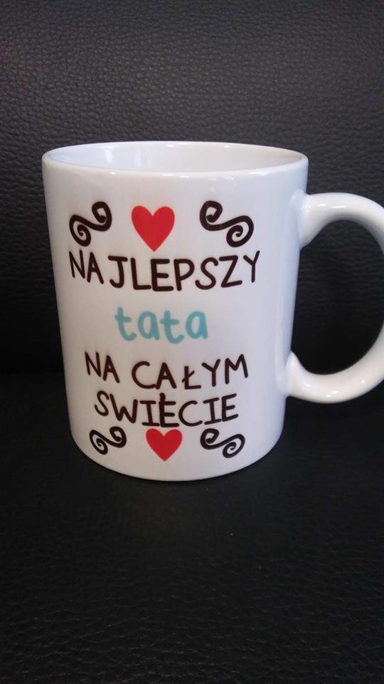 Biały kubek z napisem 'Najlepszy tata na całym świecie' w kolorach czarnym, czerwonym i błękitnym, ozdobiony zawijasami i sercami, na tle czarnej, skórzanej powierzchni.