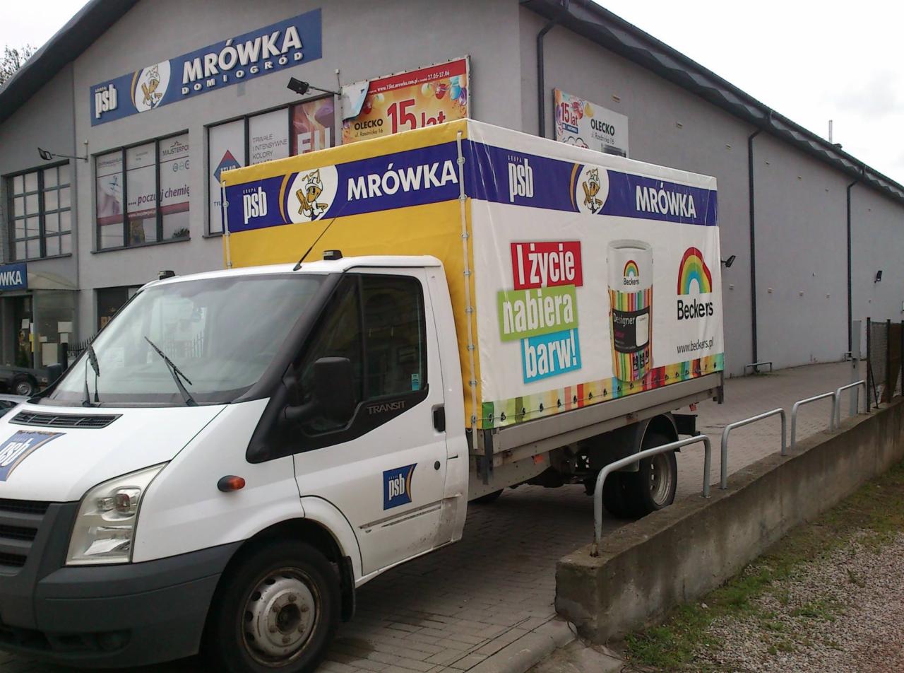 Biały samochód dostawczy z reklamą farb Beckers i logiem Mrówka na plandece, zaparkowany przed sklepem Mrówka Dom i Ogród.
