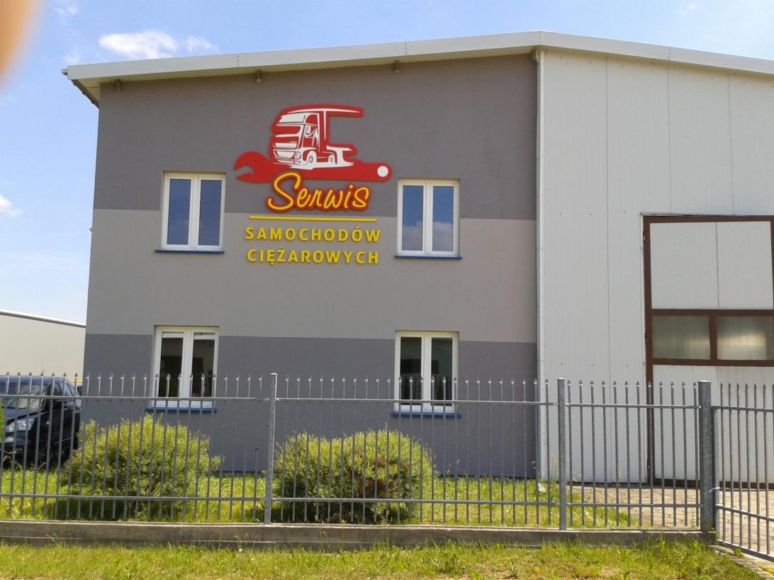 Budynek serwisu samochodów ciężarowych z logo przedstawiającym ciężarówkę w kluczu francuskim, napis 'Serwis SAMOCHODÓW CIĘŻAROWYCH' na szarej elewacji.