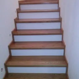 Drewniane schody z białymi podstopnicami, widok z dołu. Naturalny odcień drewna, proste wykończenie. Grzejnik na ścianie w tle.