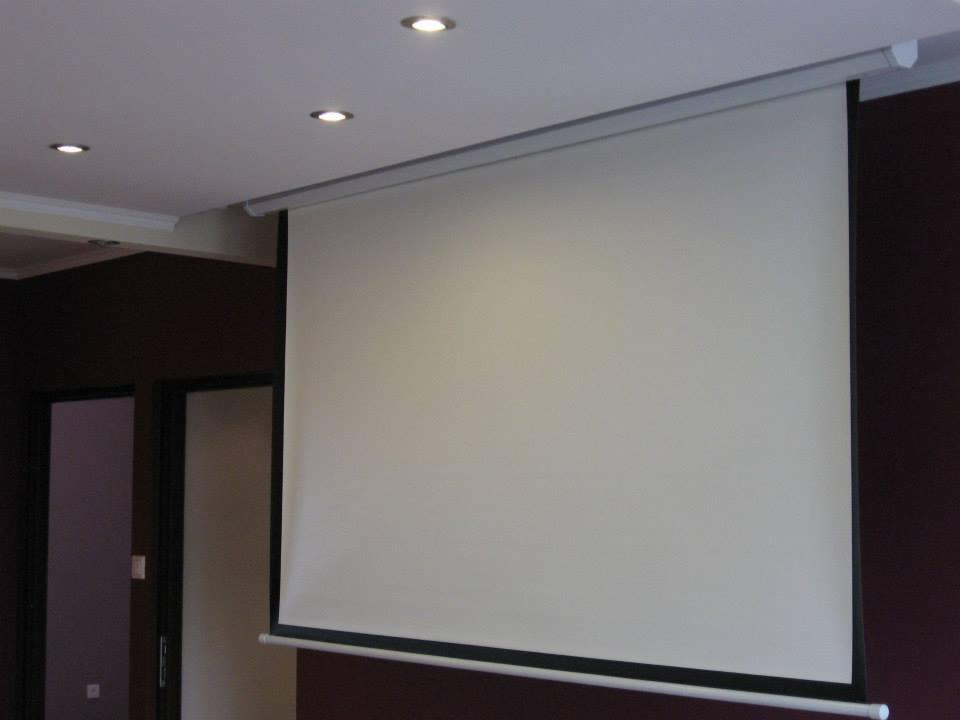 Zamontowany ekran projekcyjny zwijany, widoczne oświetlenie sufitowe punktowe i ciemne ściany.