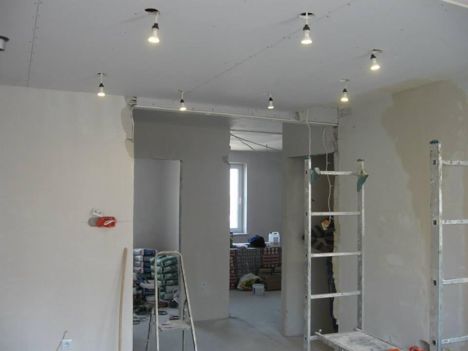Wnętrze w trakcie remontu: sufit podwieszany z oświetleniem, ściany przygotowane do malowania, drabiny malarskie, worki z zaprawą. Widok z otwartym przejściem do innego pomieszczenia.