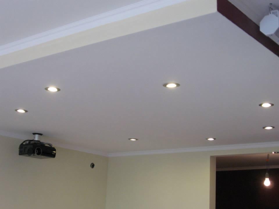 Gładki, biały sufit podwieszany z zamontowanymi punktowymi lampami LED i projektorem. Widoczne listwy dekoracyjne przy suficie i wnęka sufitowa.