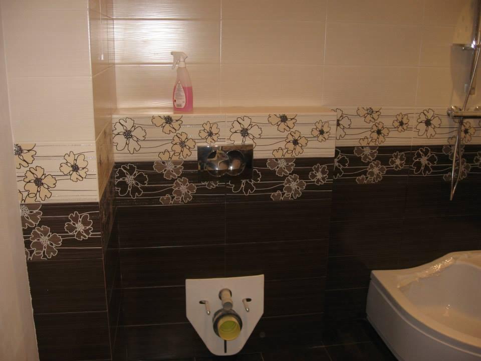 Wykończona łazienka z brązowo-białymi płytkami w kwiaty. Widoczna toaleta, fragment wanny i dozownik. Nowoczesny design z akcentem florystycznym.