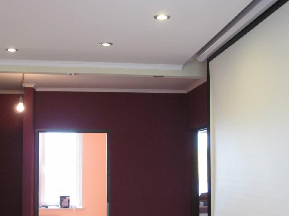 Ciemnoczerwona ściana z białym sufitem z listwami i wbudowanymi punktami świetlnymi. Widoczny fragment ekranu projekcyjnego.