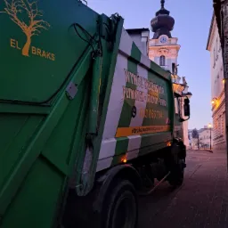 Zielona śmieciarka z logo firmy 'EL BRAKS' na tle uliczki z kościołem z wieżą zegarową w tle, wczesnym rankiem.