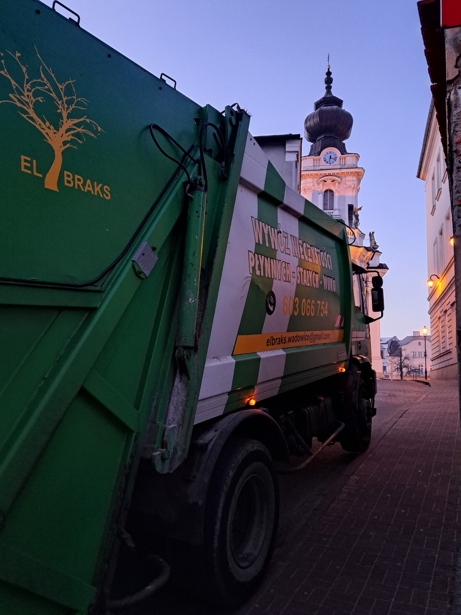Zielona śmieciarka z logo firmy 'EL BRAKS' na tle uliczki z kościołem z wieżą zegarową w tle, wczesnym rankiem.