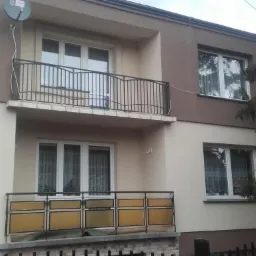 Dwukondygnacyjny dom z balkonami po remoncie elewacji, z brązowo-beżową kolorystyką, widoczną anteną satelitarną i fragmentem ogrodzenia.