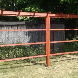 Surowa, pomalowana na czerwono drewniana konstrukcja trejażu w ogrodzie, z widocznym ogrodzeniem z siatki i zielenią w tle.