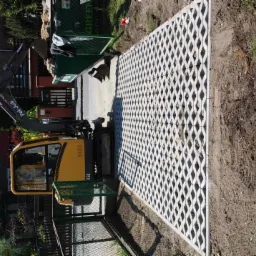 Żółta mini koparka obok ułożonych, ażurowych płyt betonowych na gruncie, widoczny budynek mieszkalny w tle.