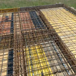 Siatka zbrojeniowa na szalunku z desek, przygotowana do wylewania betonu, widok z góry na tle zielonego pola.