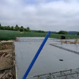 Świeżo wylana płyta fundamentowa z widocznym zbrojeniem, na tle zielonego pola i pochmurnego nieba. W kadrze widoczny fragment niebieskiego narzędzia.