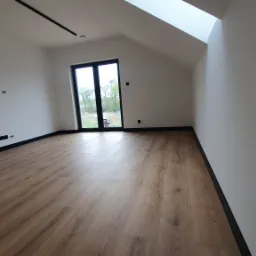 Jasne, puste poddasze z panelami podłogowymi w odcieniach brązu, świeżo pomalowane ściany na biało, czarne listwy przypodłogowe i okno dachowe wpuszczające naturalne światło.