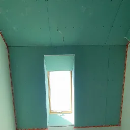 Montaż zielonej płyty gipsowo-kartonowej wokół okna dachowego, widoczne wkręty i taśmy narożnikowe. Etap prac wykończeniowych poddasza.