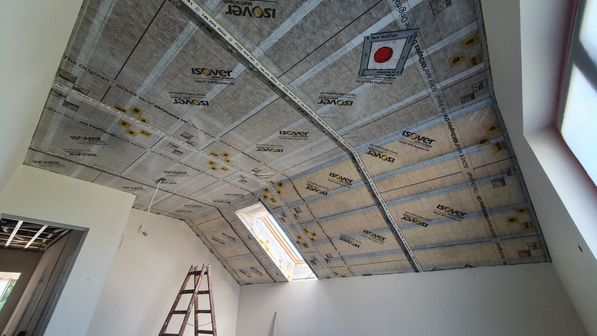 Izolacja poddasza z folią paroizolacyjną Isover Vario, widok od wewnątrz. Na skośnym suficie widoczne okno dachowe i drabina. Częściowo widoczna konstrukcja ścianki działowej.