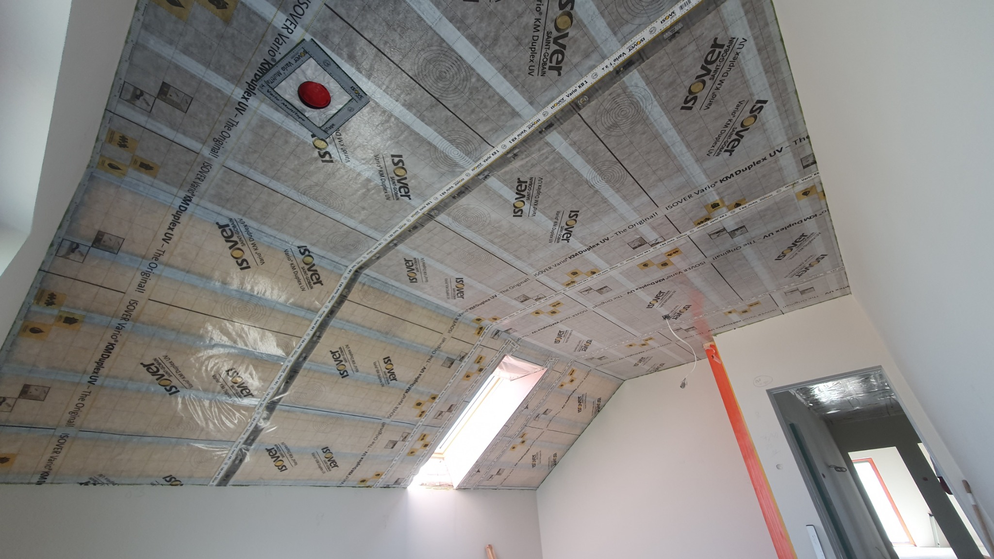 Skośny sufit poddasza z oknem połaciowym, pokryty folią paroizolacyjną Isover Vario KM Duplex UV, przygotowany do dalszych prac wykończeniowych.
